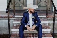 a bold rustic groom’s look with a navy suit, a white shirt and tie, a neutral hat and brown cowboy boots