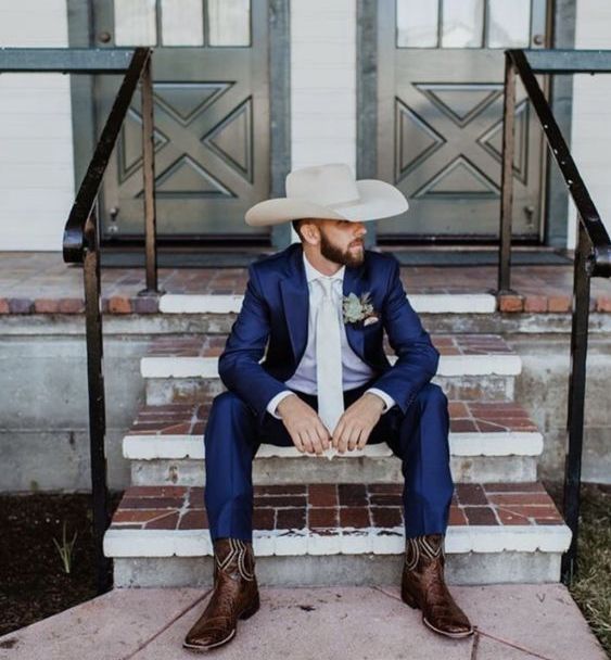 a bold rustic groom's look with a navy suit, a white shirt and tie, a neutral hat and brown cowboy boots