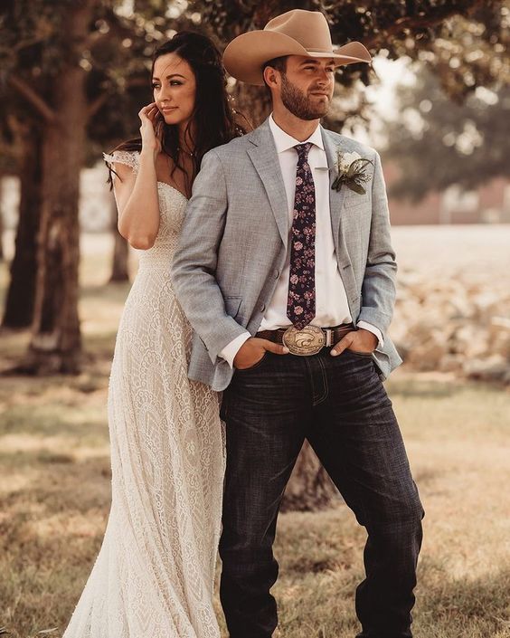 a classy rustic groom's look with a white shirt, a light blue blazer, navy jeans, a belt with a large buckle and a neutral hat