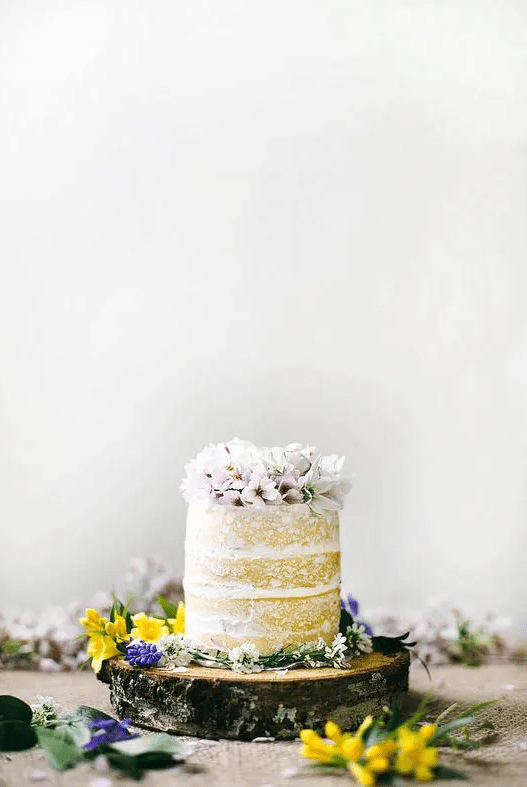 a naked wedding cake topped with fresh pink blooms and served on a wooden slice with colorful wildflowers