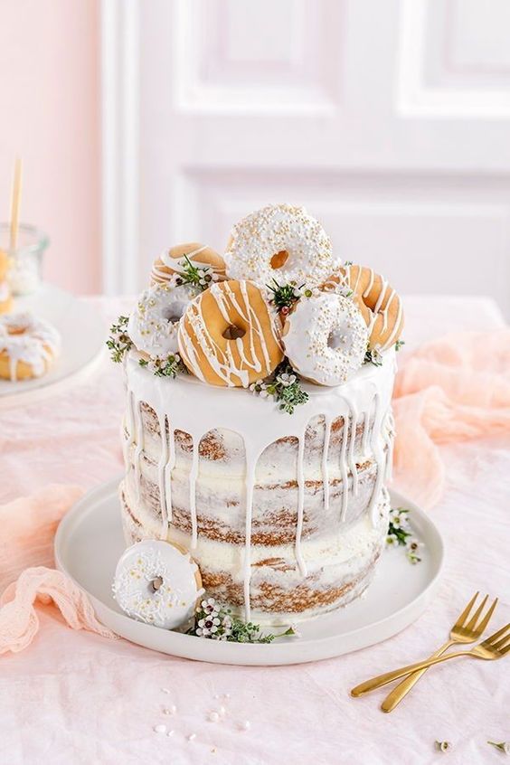 a naked wedding cake with greenery, white blooms and creamy drip plus glazed donuts on top for a spring celebration