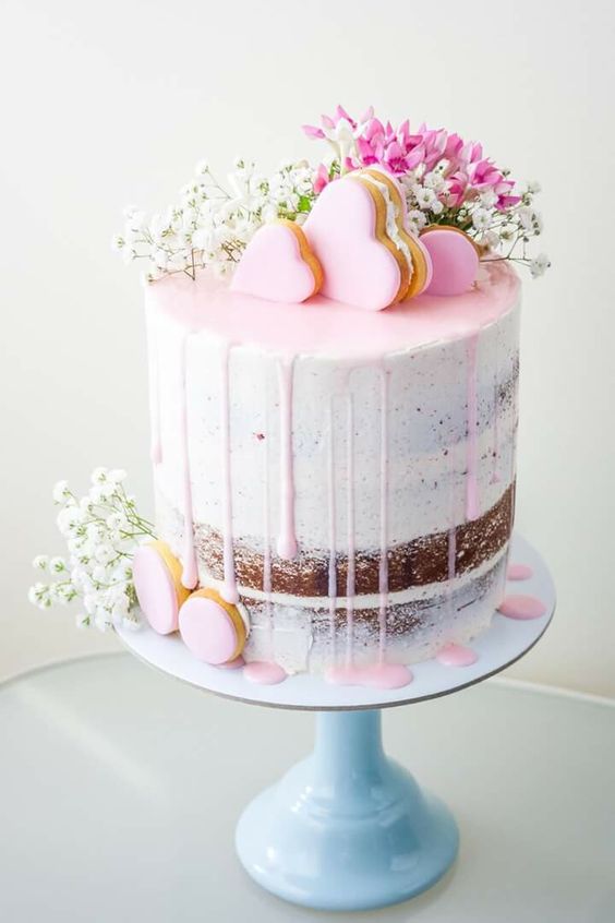 a naked wedding cake with pink drip, pink heart macarons and pink and white blooms on top the cake