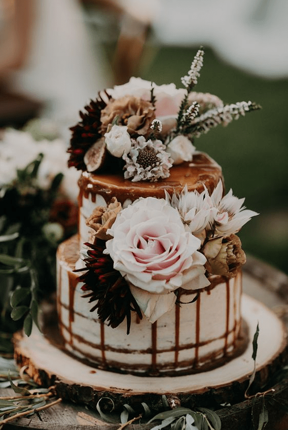 a romantic naked wedding cake with blush and burgundy blooms, with greenery and white blooms and caramel drip