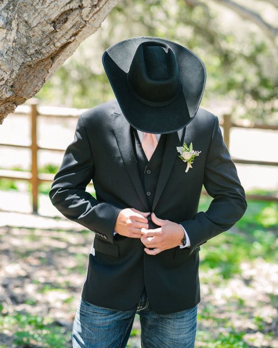 a rustic groom's look with navy jeans, a black waistcoat and a blazer, a black hat, a white shirt and a pink tie