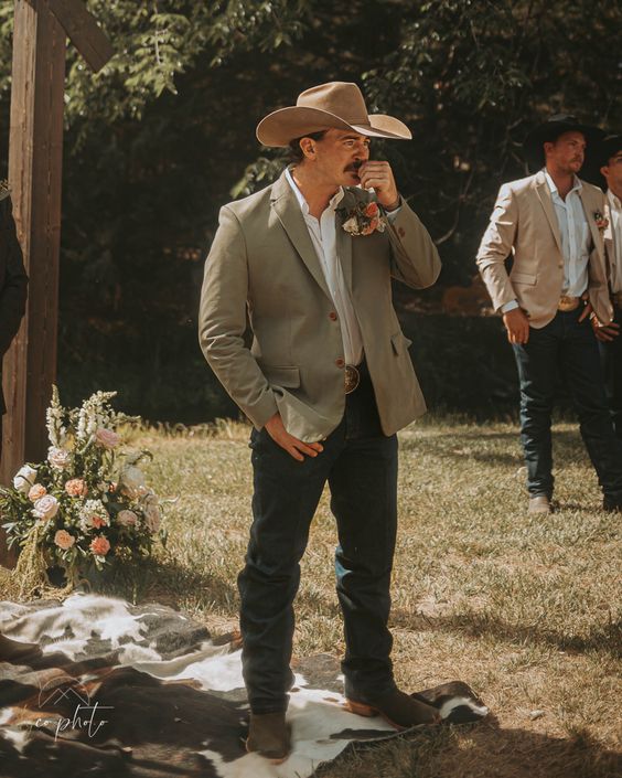 a rustic groom's outfit with a white shirt, navy jeans, brown boots, a light green blazer and a neutral hat
