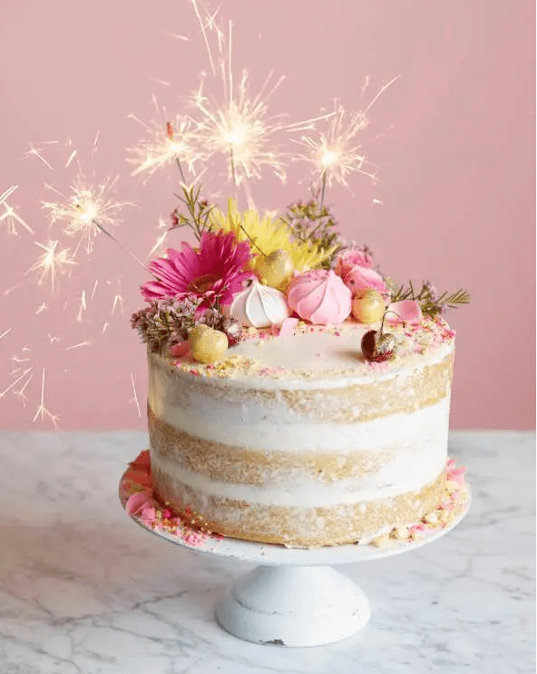 a super bright naked wedding cake with pink and yellow blooms, cherries, sparklers and bright petals and gold leaf