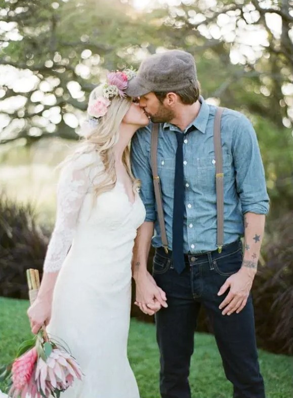 a very relaxed and rustic groom's outfit with navy jeans, a blue chambray shirt, a navy tie, suspenders and a cap