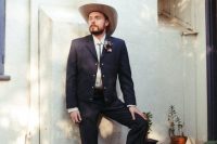 a western groom’s look with a black suit, a white shirt, a bolo tie, a neutral hat and brown cowboy boots