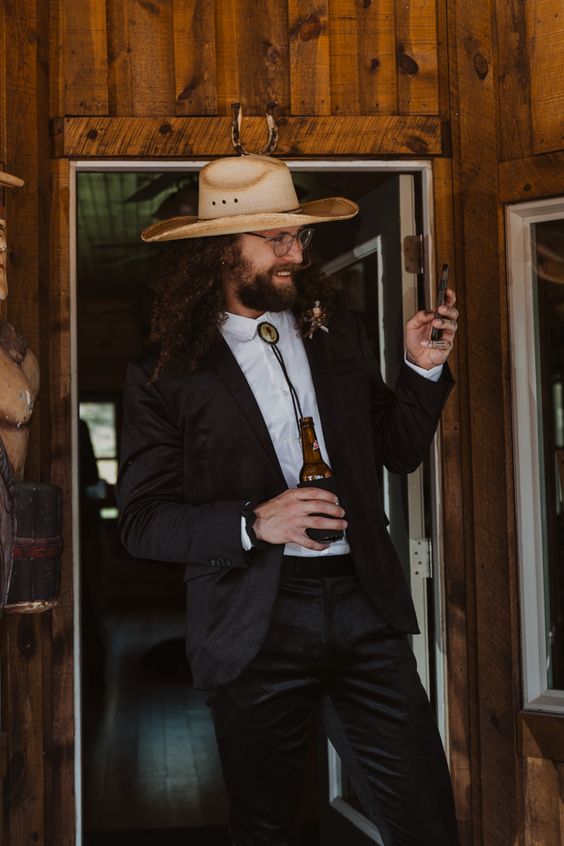 a western groom's outfit with a black suit, a white shirt, a bolo tie and a neutral hat is a cool idea for a boho wedding, too