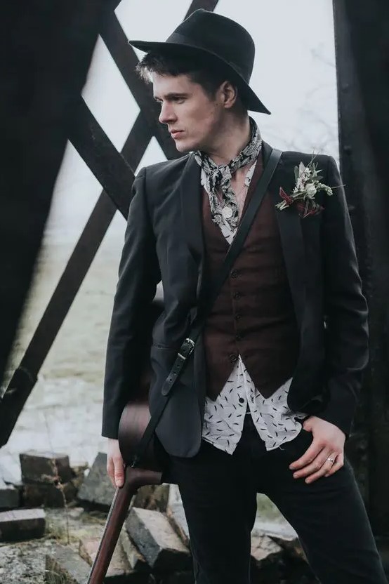 an alternative groom's look with a printed shirt, a brown waistcoat, a black blazer, a neck tie, a black hat and a boutonniere