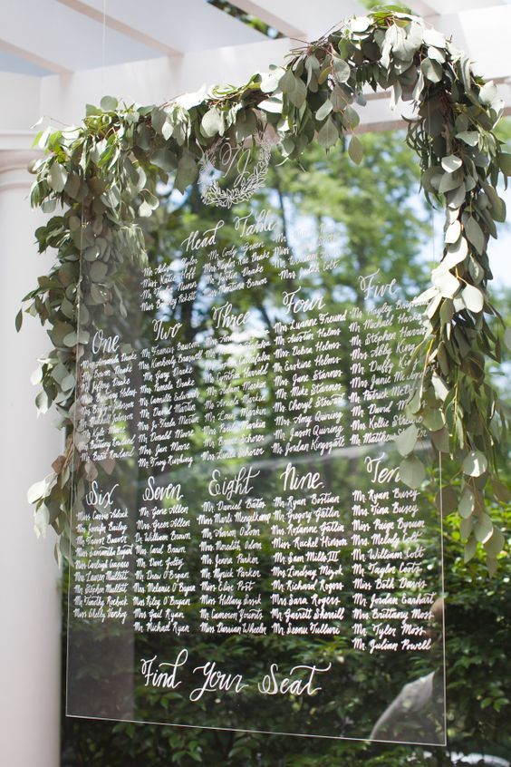 a hanging acrylic seating chart topped with a fresh greenery garland