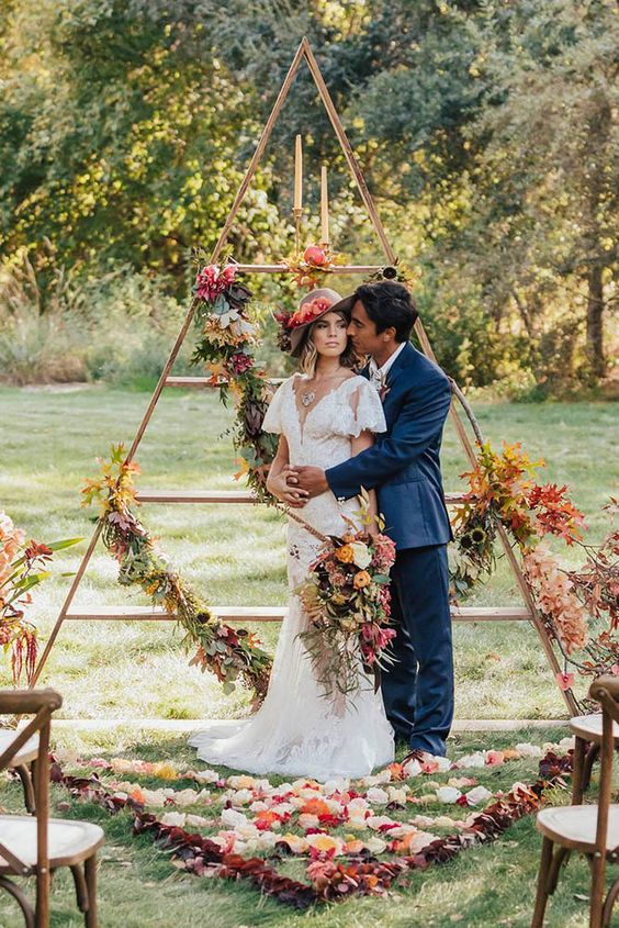 a tall triangle wedding altar with fall leaves, lush blooms and candles for a fall wedding