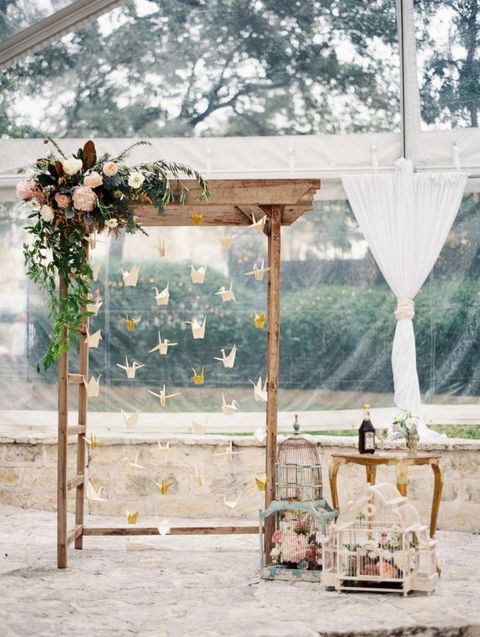 a wedding arch with lush greenery and blooms and hanging paper cranes, bird cages and blooms