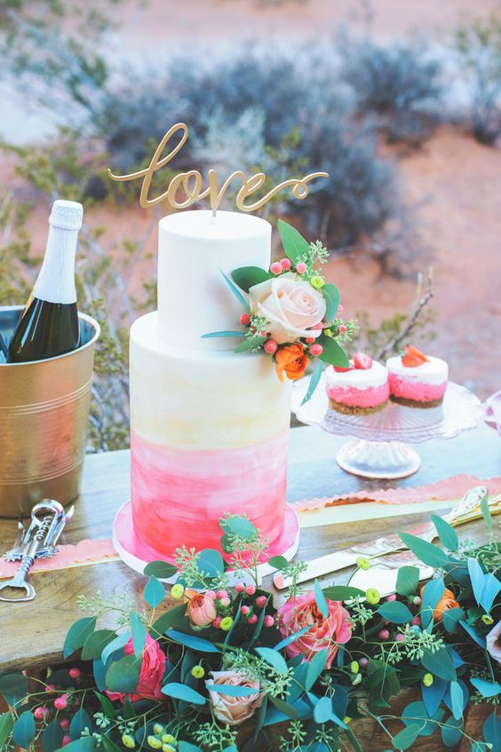 a coral and pink watercolor wedding cake is topped with a flirty gold cake topper