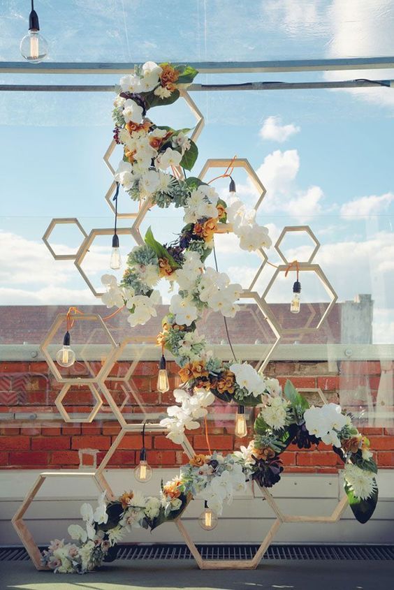a wooden honeycomb wedding backdrop with bulbs, lush blooms and greenery for a trendy geo touch