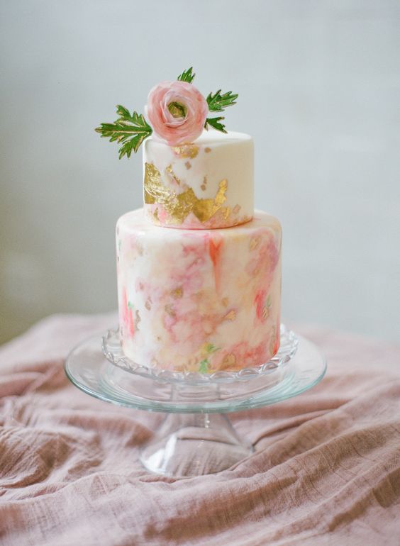 a pink and coral gilded watercolor wedding cake with a pink bloom and some leaves on top