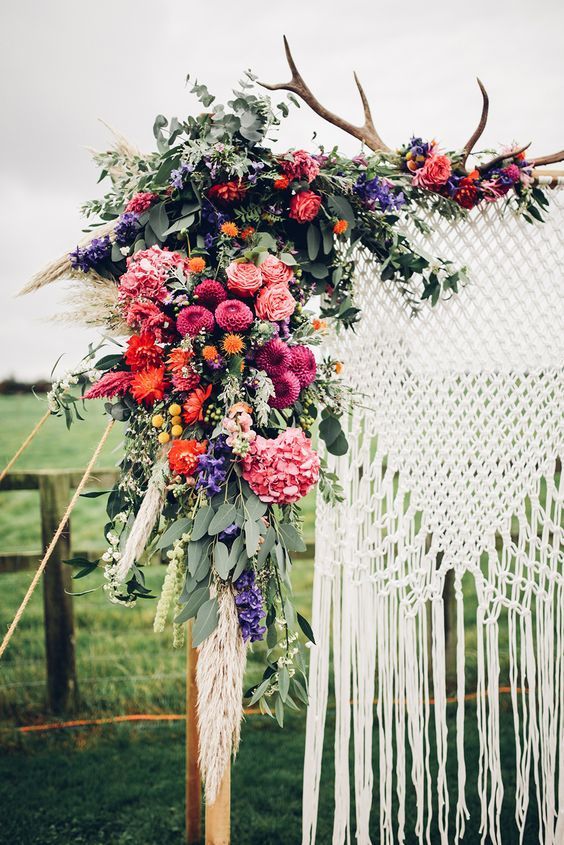 a lush colorful floral wedding arch with antlers and macrame hanging for a boho wedding
