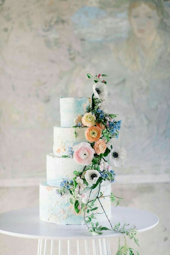a light blue wedding cake with sugar watercolor blooms painted and real flowers and greenery attached on one side