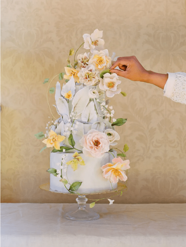 a pale blue wedding cake decorated with a lot of yellow, blush and pale blue flowers and greenery