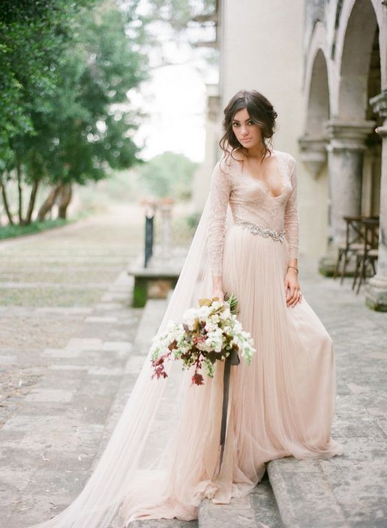 a blush wedding dress with a V neckline, long sleeves, an embellished belt and a matching veil