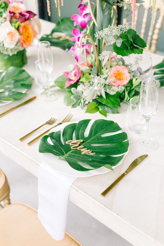 a tropical table setting with lush leave sand flowers plus gold flatware for a bridal shower
