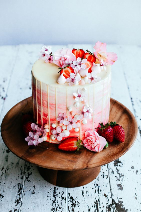 a tender watercolor pink and coral wedding cake with cream drip, cherry blossom and cut strawberries on top and sides