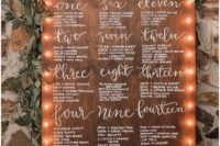 23 a wooden seating chart with white calligraphy and marquee lights and topped with fresh greenery and blush blooms