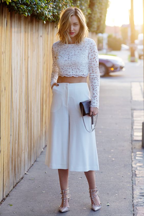 a white lace crop top with long sleeves, cropped culottes, blush studded shoes and a black clutch