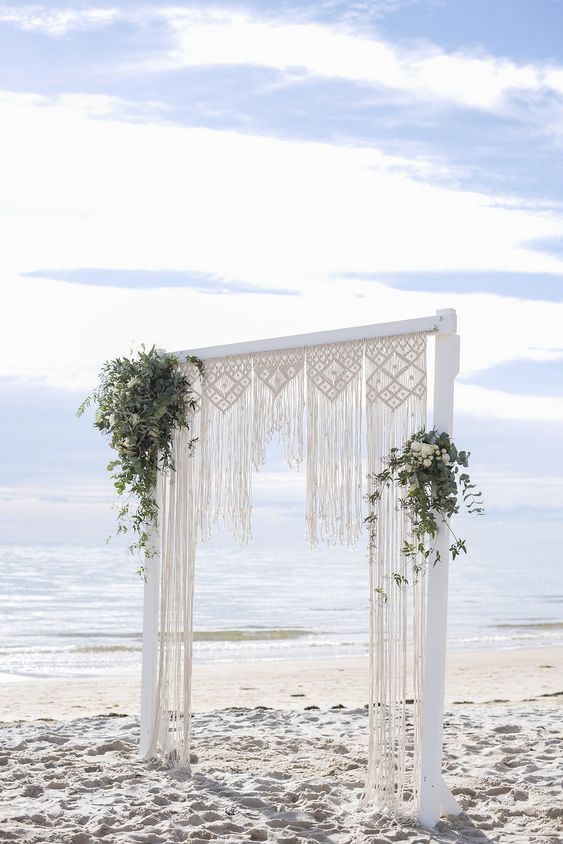 a marcrame wedding arch with greenery and white blooms for a beach boho wedding