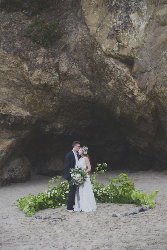 a lush greenery wedding arch with some white blooms and pebbles right on the beach