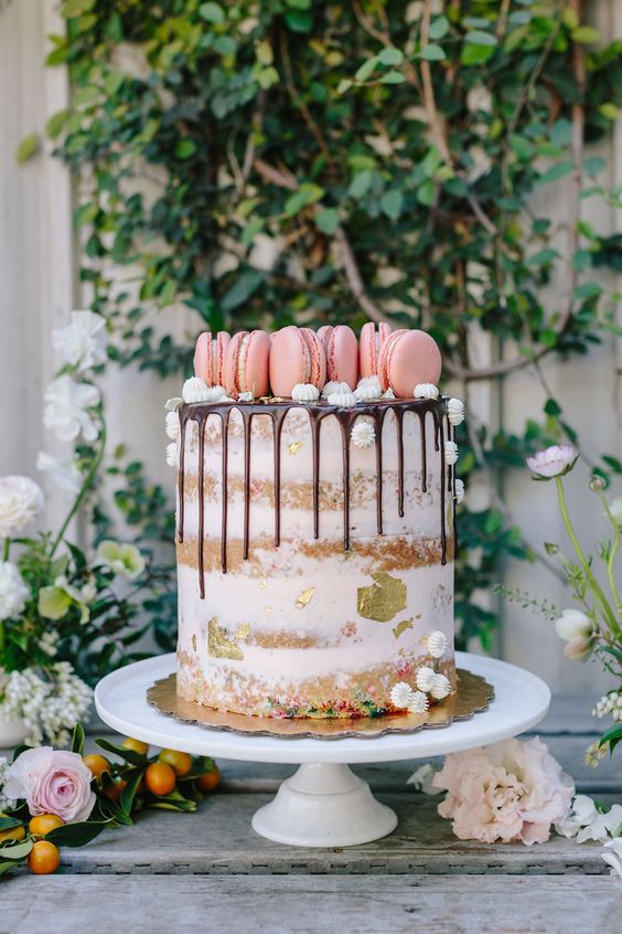 a semi-naked cake with gold foil and topped with pink macarons on top plus small meringues