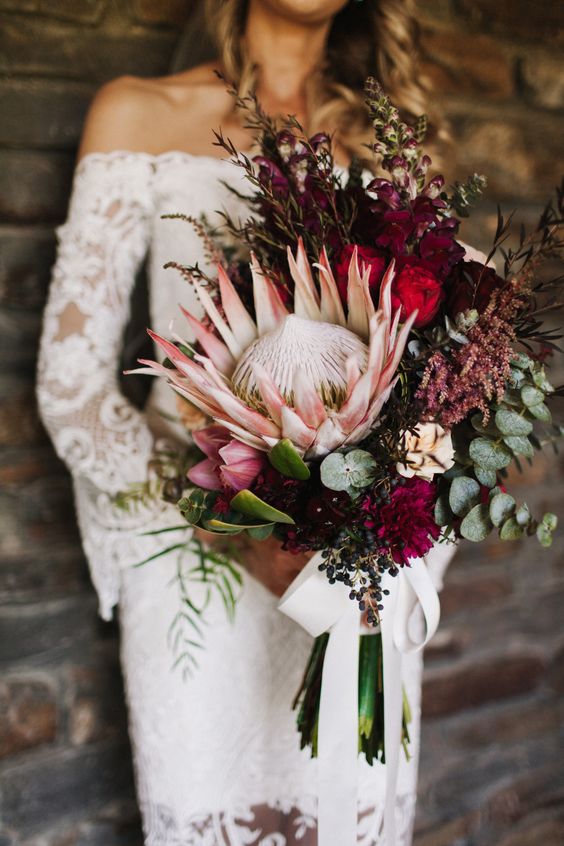 a fall boho wedding bouquet with a vertical shape, a king protea, burgundy and plum blooms, herbs and greenery plus a bow