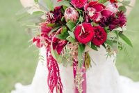 a berry-hued wedding bouquet of red and fuchsia roses, lilies, king proteas, greenery, amaranthus and long ribbon for a fall wedding