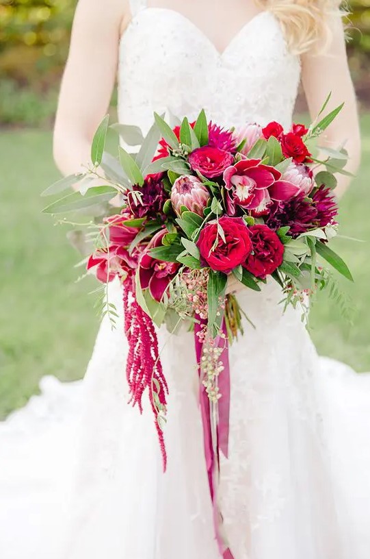a berry-hued wedding bouquet of red and fuchsia roses, lilies, king proteas, greenery, amaranthus and long ribbon for a fall wedding