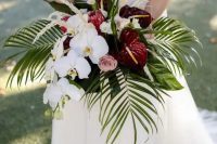 a tropical wedding bouquet of white orchids, blush roses, king protea and burgundy anthurium and leaves