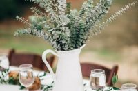 a white milk jug with eucalyptus, a greenery table runner and dark candle holders with candles