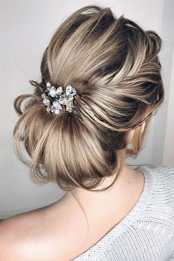 a messy and textural low chignon hairstyle with a light side braid and a large rhinestone and pearl hairpiece