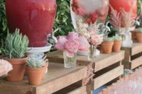 a pretty and creative drink bar with crates as stands, potted greenery and succulents, blooms, pink glasses is a very stylish idea