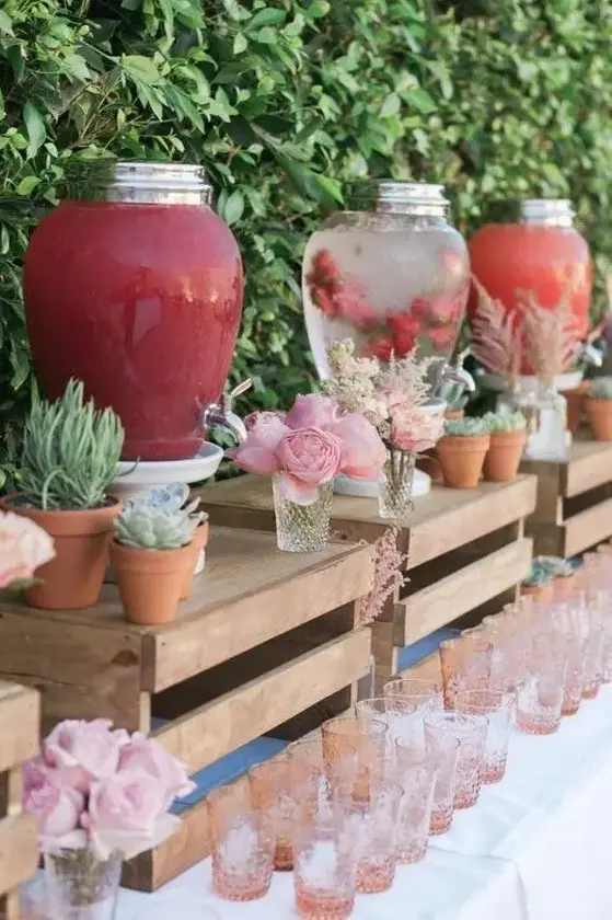 a pretty and creative drink bar with crates as stands, potted greenery and succulents, blooms, pink glasses is a very stylish idea