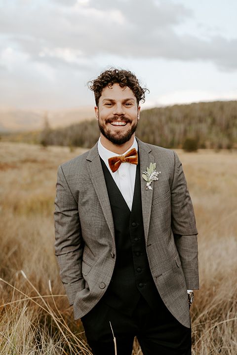 a rustic groom's outfit with a grey blazer, a black waistcoat and pants, a white shirt and a rust velvet bow tie