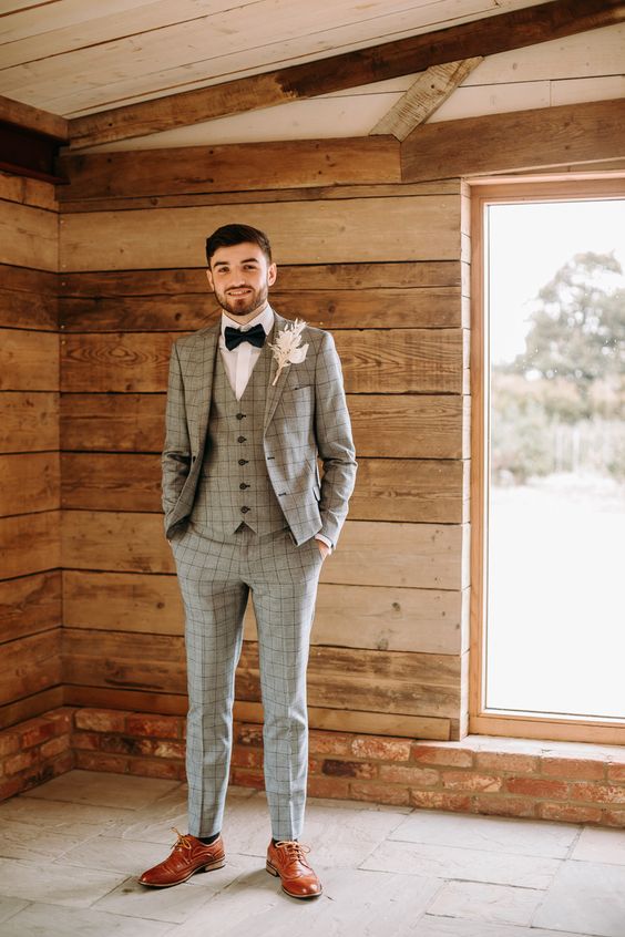 a stylish grey three-piece windowpane suit, a white shirt, a black bow tie and amber shoes plus a dried flower boutonniere