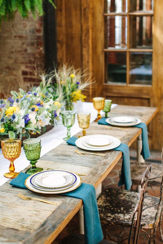 a chic rustic setting with colored glasses, blue napkins and a lush floral centerpiece with thistles