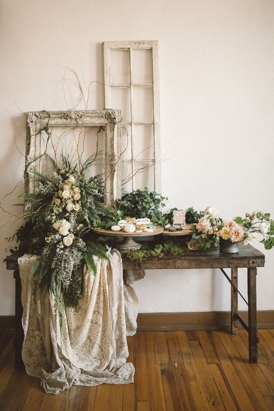 a rustic garden wedding dessert table with plenty of greenery and blooms and vintage frames plus lace