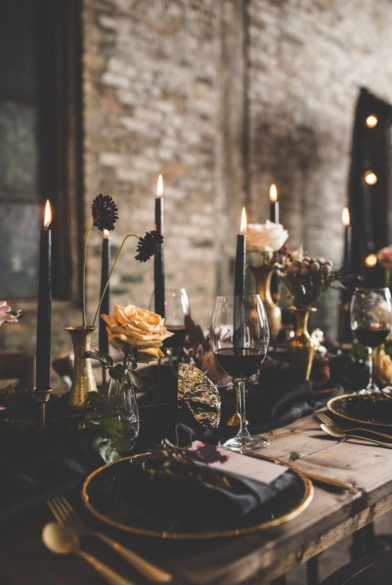 a raw wooden table with a black table runner and black candles, gilded touches and moody blooms