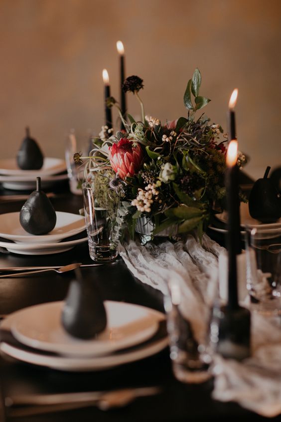 an elegant moody tablescape with a textural and herb centerpiece, black candles and pears and a black table