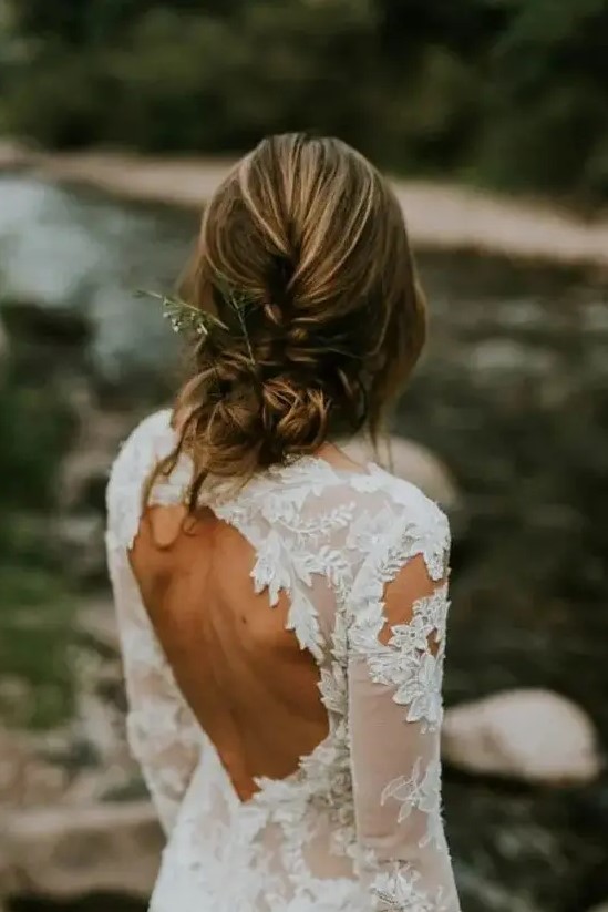 a casual braided and twisted low updo with bangs with some greenery in it looks a bit messy and it makes it great for a destination wedding