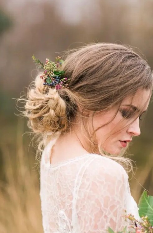 a low updo with a braided and twisted low bun, a volume on top, some locks and some fresh greenery and blooms for a boho wedding