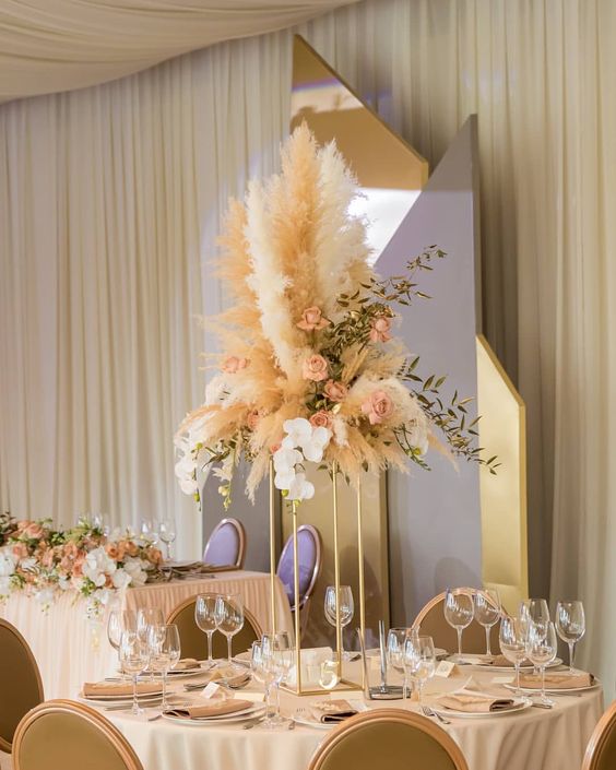 a creative wedding centerpiece of dusty pink roses, greenery and pampas grass on a tall gold stand