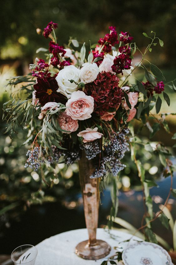 a lush floral centerpiece of burgundy, blush and white blooms, textural greenery in a gold vase