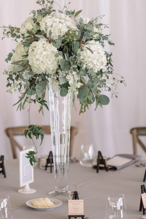 a lush floral centerpiece of eucalyptus and white hydrangeas in a tall glass vase